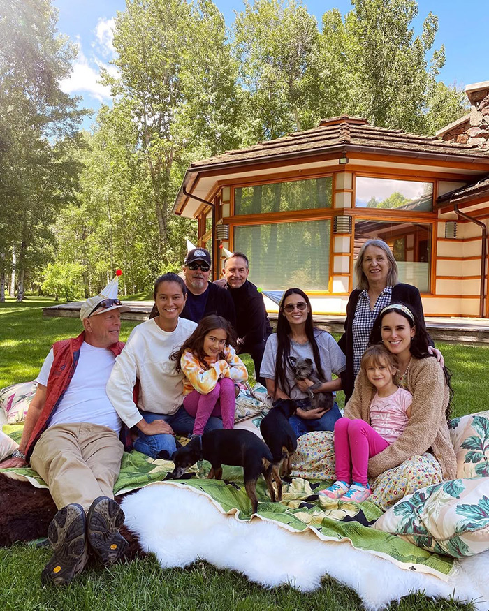Family gathering on a lawn, including Bruce Willis’ wife, a house in the background, and dogs playing nearby.