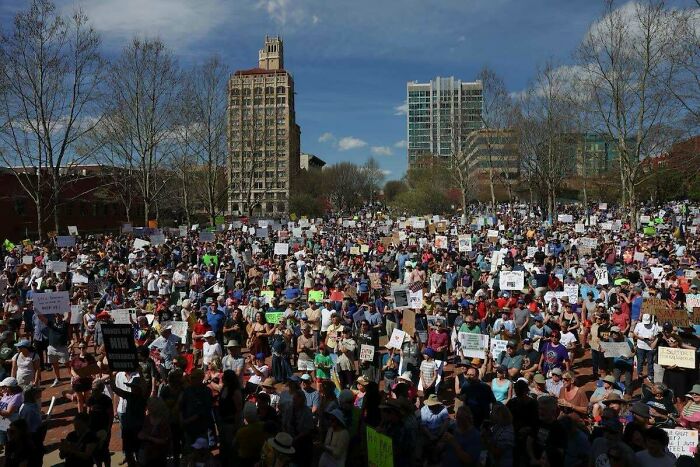 Large chaotic good crowd gathered outdoors in a city plaza holding signs and protesting on a sunny day.