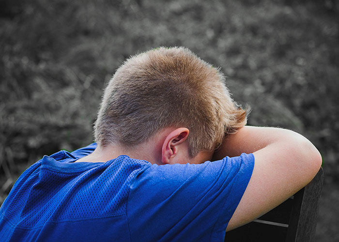 Boy wearing a blue shirt resting his head on his arms, depicting a take one for the team moment in an outdoor setting.